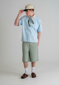 Light blue oversized t-shirt, green denim shorts, beige cap, and patterned scarf, paired with brown clogs and white socks.