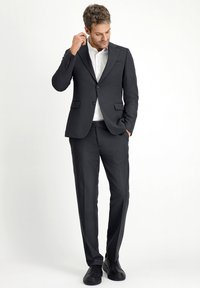 Suit nero con rever a punta, due tasche frontali e pantaloni abbinati. Indossato sopra una camicia bianca e abbinato a scarpe nere.