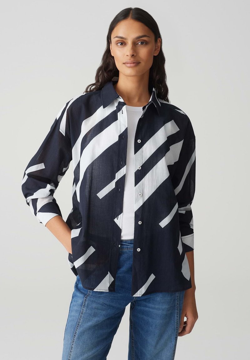 Camicia blu navy con bottoni, a strisce diagonali bianche, in tessuto leggero, maniche lunghe arrotolate fino ai gomiti e una vestibilità rilassata.