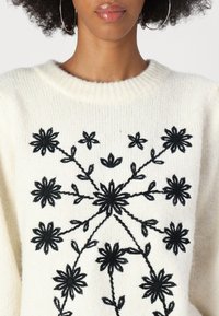 Maglione crema con texture a maglia, caratterizzato da ricami floreali neri in un motivo circolare sul petto e maniche lunghe e rilassate.