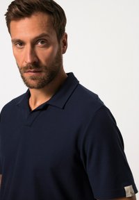 Camisa polo azul marino hecha de tela texturizada, con cuello clásico y mangas cortas. Detalle notable en la manga con una etiqueta de logo.