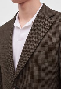 Homme portant un blazer marron à motif pied-de-poule sur une chemise blanche à col, montrant la partie supérieure de la poitrine et du cou.