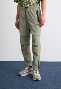 Light olive green Nike joggers with an adjustable waistband, zippered side pocket, and black accents. Paired with gray running shoes.