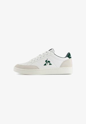 Baskets basses blanches avec un empiècement en daim beige au niveau de la pointe et du talon, logo vert foncé et étiquette de talon, et lacets blancs sur fond blanc.