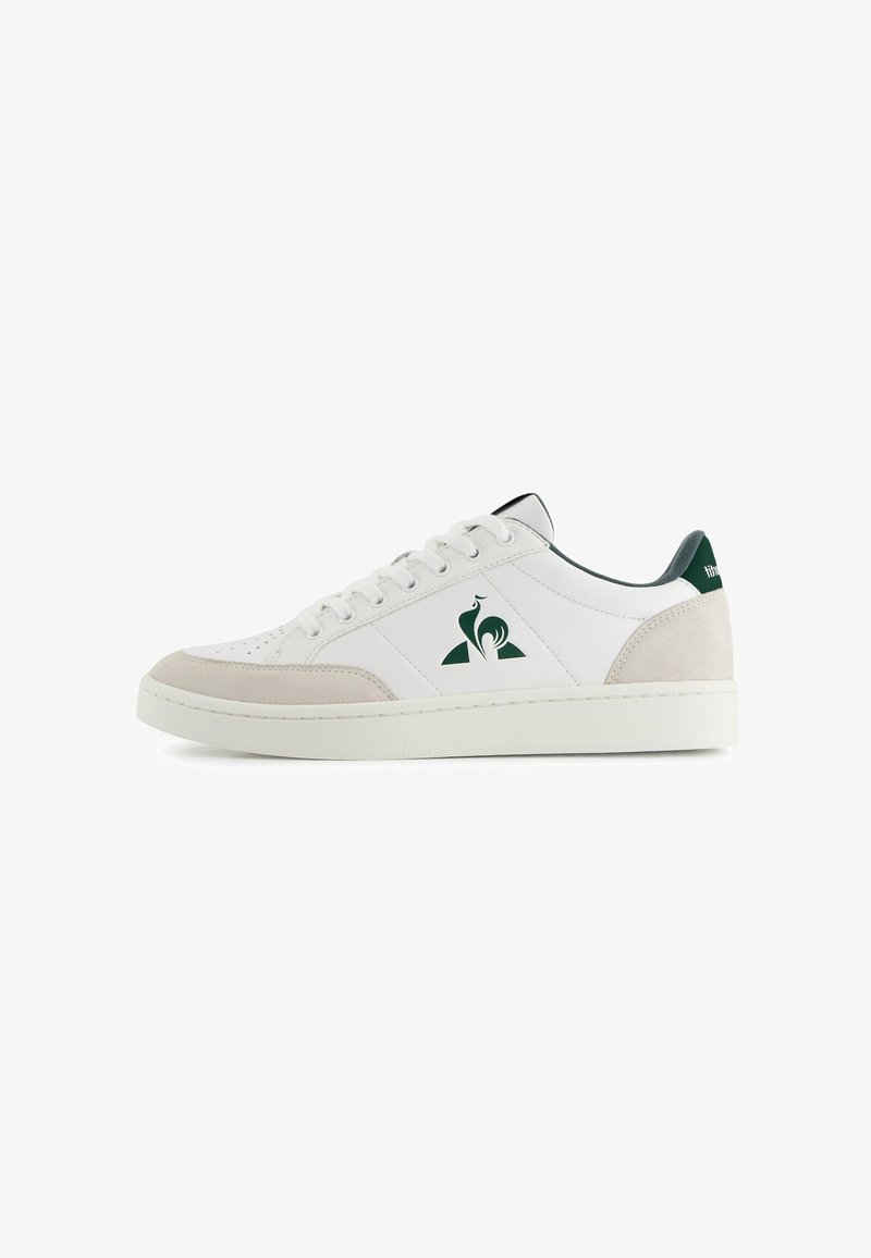 Zapatilla baja blanca con punta y talón de ante beige, logotipo verde oscuro y tira del talón, y cordones blancos sobre un fondo blanco.