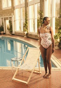 Rožinė, gėlėmis dekoruota vientisa maudymosi kostiumėlį su reguliuojamais petnešomis. Medinė poilsio kėdė su eskiziniais piešiniais šalia baseino.