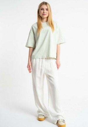 Młoda kobieta z długimi blond włosami stoi ubrana w luźną, jasnooliwkową koszulkę, szerokie białe spodnie i beżowe sneakersy na platformie.