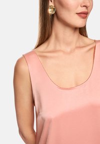 Rosa Satintop mit einem weiten Rundhalsausschnitt, verziert mit feinen goldenen Nähten am Rand, ergänzt durch große goldene kreisförmige Ohrringe.