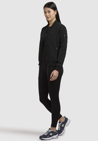 Svart bomberjacka har dragkedja framtill, ribbade ärmslut och två sidofickor. Kombinerad med svarta leggings och blåa träningsskor.