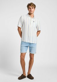 Camicia a maniche corte a righe blu chiaro con patta dei bottoni, abbinata a pantaloni corti in denim chiaro, indossata con zoccoli marroni. Tessuto dalla texture liscia.