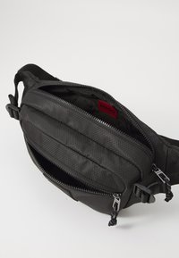 Schwarze strukturierte Stoff-Waistbag mit zwei Reißverschlussfächern, sichtbares rotes HUGO-Logo im Hauptfach.