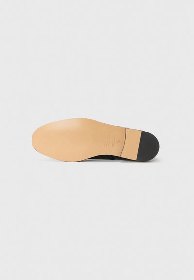 Sole di scarpa in pelle nera con fondo chiaro, caratterizzata da una superficie liscia e una forma arrotondata. Presenta un marchio in rilievo e dettagli vicino al tallone.