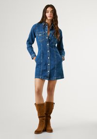 Robe chemise en denim bleu avec manches longues, poches poitrine, boutonnière devant et ourlet évasé. Associée à des bottes en suède marron arrivant au genou.