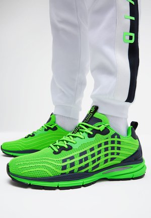 Helle grüne und schwarze Sportschuhe mit gemusterten Mesh-Seiten, getragen zu weißen Hosen mit einem schwarzen und grünen Seitenstreifen.