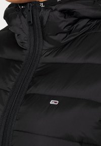 Black puffer jacket with zip front, high collar, and horizontal stitching. Features a small logo patch on the chest. Smooth texture.