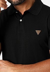 Polo negro hecho de tela suave, con un collar clásico, tapeta de tres botones y un logo triangular marrón en el pecho.