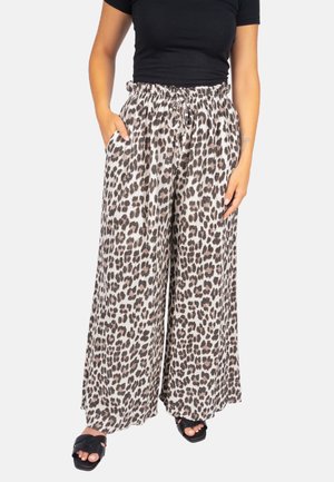 Frau trägt weite Leopardenmuster-Hosen mit elastischem Bund und schwarzem Oberteil, steht mit teilweise in die Taschen gesteckten Händen und schwarzen Sandalen.