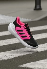 Black athletic shoe with pink accents, featuring a mesh upper, white sole, and three black stripes. Design emphasizes lightweight and breathable materials.