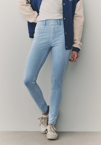 Svetlo modre ozke kavbojke z visokim pasom, s gladko teksturo in minimalnimi šivi, kombinirane z bež supergami.