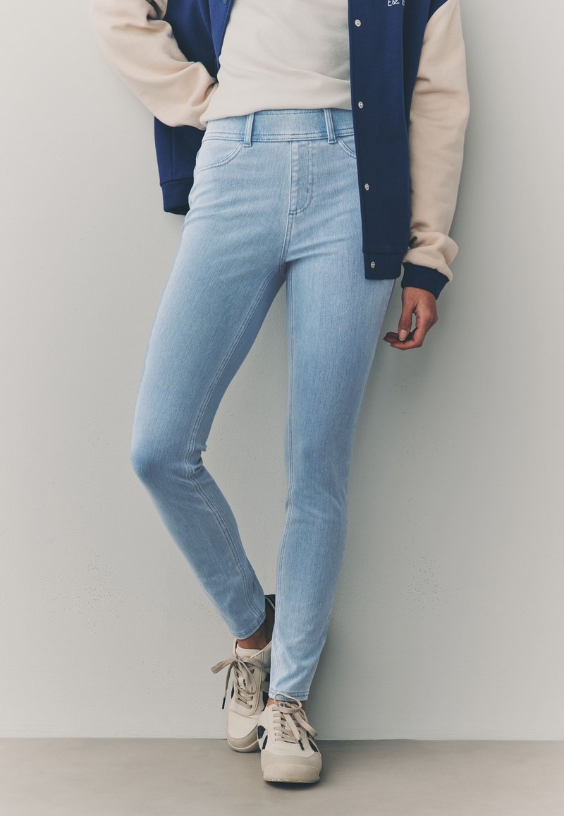 Svetlo modre ozke kavbojke z visokim pasom, s gladko teksturo in minimalnimi šivi, kombinirane z bež supergami.