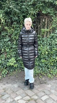 Črn podložen dolgi plašč z pasom, kapuco in sijočo teksturo. Nosi se čez svetlo modre hlače in črne čevlje, postavljen ob zeleni vegetaciji.