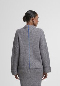 Grau gestrickter Pullover mit hohem Kragen, gerippter Textur und einem zentralen blauen Streifen auf der Rückseite. Die Ärmel sind weit mit gerippten Bündchen.