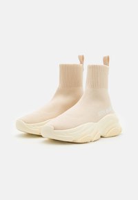 Baskets à chaussettes beige tricotées jusqu'à la cheville avec une tige texturée, une languette et une semelle en caoutchouc épaisse présentant un design ondulé et une marque blanche.