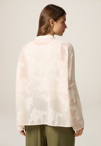 Blusa leggera e trasparente color crema con motivo floreale testurizzato; presenta maniche lunghe e un colletto rotondo, indossata sopra pantaloni verde oliva.