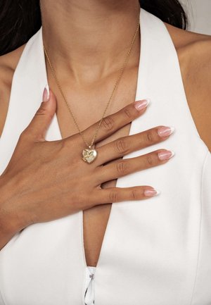 Hand with manicured nails touching chest, wearing white sleeveless top and gold heart-shaped locket necklace with butterfly detail.