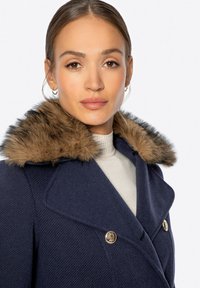 Marineblauer Wollmantel mit einem Kunstfellkragen, doppelreihigem Design und goldfarbenen Knopfakzenten, über einem hellgrauen Oberteil getragen.