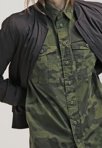 Schwarze Bomberjacke über einem grünen Camouflage-Hemd mit Knopfleiste, zwei Fronttaschen und Kragen. Die Jacke hat eine glatte Textur.