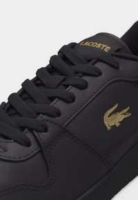 Μαύρο αθλητικό παπούτσι Lacoste με μαύρα κορδόνια, χρυσό κεντημένο λογότυπο κροκόδειλου στο πλάι και χρυσή επωνυμία Lacoste στη γλώσσα.