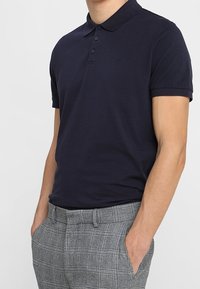 Polo shirt bleu marine avec un col, trois boutons et un petit logo. Porté avec un pantalon en tartan gris avec un devant plat et des poches.