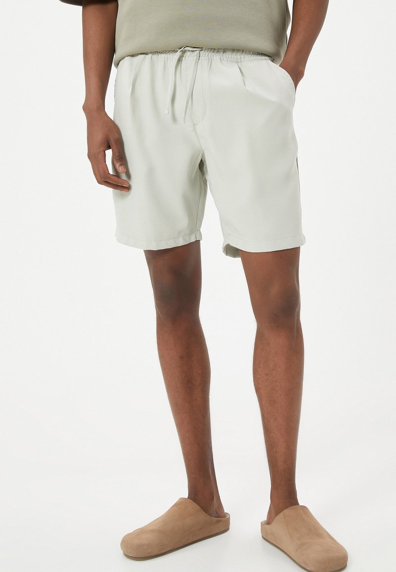 Pantalones cortos de color gris claro con cintura elástica y cordón, que presentan un ajuste holgado y una textura suave, combinados con zapatos de lona beige.