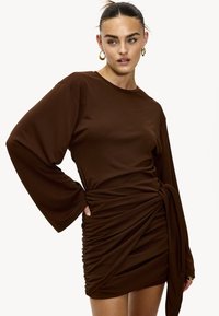 Robe marron à manches longues avec une coupe ample, des fronces sur le côté et un détail noué à la taille, confectionnée dans un tissu doux.