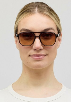 Blonde Frau mit dunkelbrauner, übergroßer Sonnenbrille mit Verlaufsgläsern, weißem Oberteil, neutralem Hintergrund, leichtem Lächeln und Blick nach vorne.