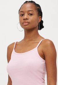 Lys rosa tank topp med tynne stropper og rund hals. Glatt tekstur, tettsittende design og minimalt med sømdetaljer. Store hoop øredobber.