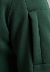 Green sweatshirt with a textured, ribbed fabric. Features a padded shoulder area and a side pocket with a concealed zipper.