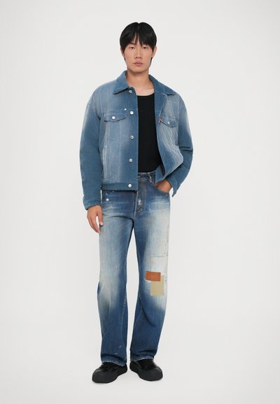Young man wearing faded blue denim jacket, black shirt, patched blue jeans, and black sneakers standing against white background.