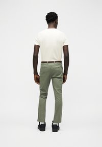 Άνδρας π ου στέκεται με την πλάτη στραμμένη φορώντας λευκό πουκάμισο με κοντά μανίκια, χακί παντελόνι με καφέ ζώνη, λευκές κάλτσες και μαύρα αθλητικά παπούτσια.