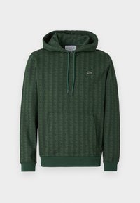 Hanorac verde cu un model geometric, glugă cu șnur și manșete ribbate. Logo-ul Lacoste de culoare contrastantă pe piept.