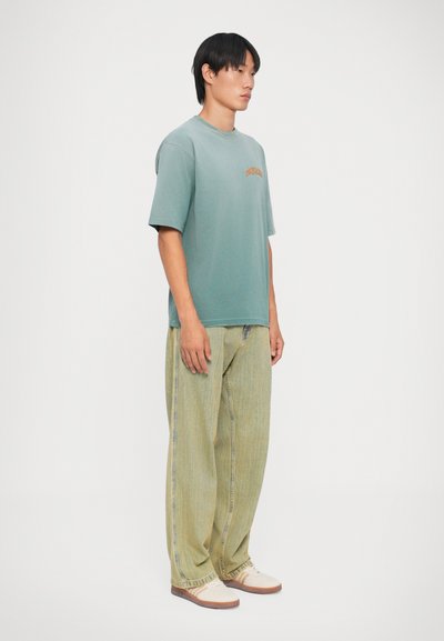Mintgroene oversized t-shirt met korte mouwen en een logo aan de voorkant, gecombineerd met lichtgroene gestreepte wijde jeans en beige schoenen.