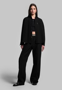 Zwarte oversized shirt met een knoopsluiting, gecombineerd met hoge zwarte broeken. Glad textuur, minimale branding en een relaxed fit.