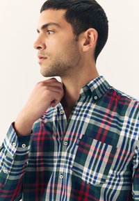 Chemise à carreaux multicolores avec des motifs verts, rouges, bleu marine et blancs, dotée d'un col boutonné et d'une poche unique sur la poitrine.