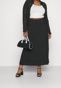 Zwarte maxi rok van vloeiend materiaal, gecombineerd met een witte tanktop en een doorschijnende zwarte blouse met knopen. Accessoires zijn een kleine zwarte handtas met witte accenten.