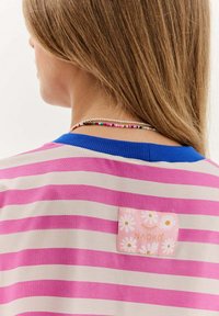 Pruhovaná košile s krátkým rukávem v světle béžové a růžové barvě, s modrým žebrovaným límečkem; obsahuje čtvercovou růžovou cedulku s květinovým vzorem a názvem značky.