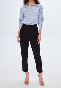 Femme portant une chemise boutonnée bleu rayé rentrée dans un pantalon noir taille haute et des sandales à talons beiges avec des lanières.