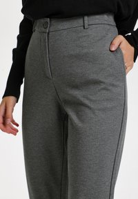 Personne portant un pantalon gris taille haute avec bouton et fermeture éclair, une main dans la poche, un haut noir à manches longues visible à la taille.