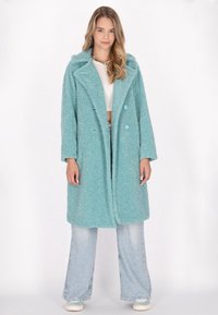 Cappotto turchese e soffice con colletto a revers, due grandi bottoni blu e tasche laterali. Abbinato a jeans in denim chiaro a gamba larga.