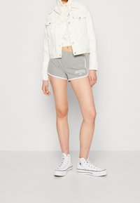 Žena v bílém zkráceném džínovém džinovém saku, bílé mikině s kapucí, šedých šortkách Hollister a bílých platformových kotníčkových teniskách stojící na jednobarevném pozadí.